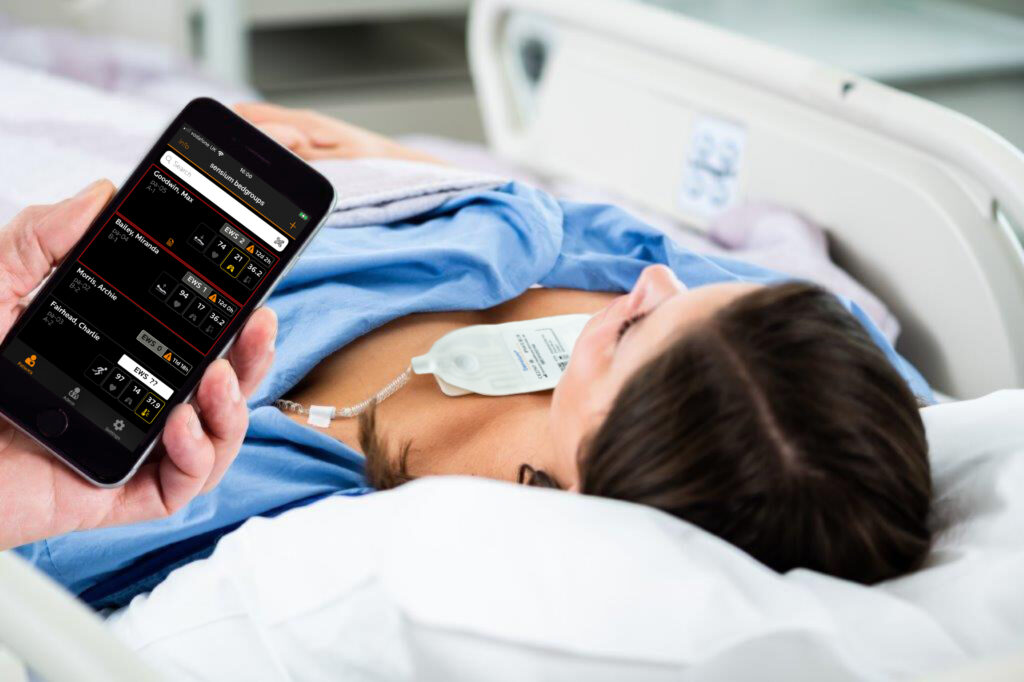 Doctor holds mobile phone with Sensium app open showing patients vitals and EWS, female patient with Sensium patch on hospital bed